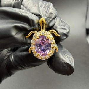 Op de afbeelding: Goudkleurige schildpendant met een grote, ovaal geslepen paarse edelsteen in het midden, omringd door kleinere, heldere stenen. De schildpadschild is geaccentueerd met heldere stenen en de kop en vinnen zijn goudkleurig. Een stijlvol sieraad.
