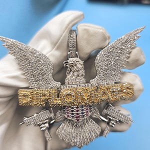 May include: A diamond-studded pendant in the shape of an eagle with wings spread wide. The word "DIPLOMATS" is written in gold. Two small guns are visible below the wings. The pendant is held by a gloved hand.