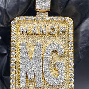 May include: A rectangular gold pendant with the words "MAN OF GOD" and the initials "MG" encrusted with sparkling white gemstones. The pendant has a gold bail and is set against a dark background.