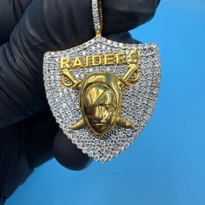 May include: A gold-toned pendant featuring the word "RAIDERS" and a stylized face with crossed swords. The pendant is encrusted with small, clear stones, and is shield-shaped. The bail is also encrusted with small, clear stones.