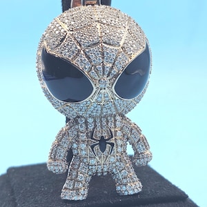 May include: A gold-coloured pendant depicting a stylized Spider-Man figure, covered in small, glittering stones. The figure features large black eyes and a spider emblem on the chest. The pendant is suspended from a black clasp.