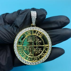 May include: Gold-coloured pendant with a circular design, featuring the words "LOVE," "LIFE," and "LOYALTY." The pendant is encrusted with small, sparkling stones around the edge and on the bail. The background is a solid light blue.