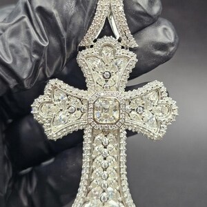 May include: A large, ornate pendant in the shape of a cross, encrusted with numerous clear, sparkling gemstones. The pendant features a central square stone surrounded by smaller gems, with intricate detailing throughout. The piece is held against a dark background.