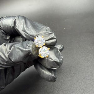 Peut inclure: Deux boucles d'oreilles à clous dorées avec de grosses pierres rondes et transparentes. Les boucles d'oreilles sont tenues par une main gantée sur un fond sombre. Les pierres scintillent.