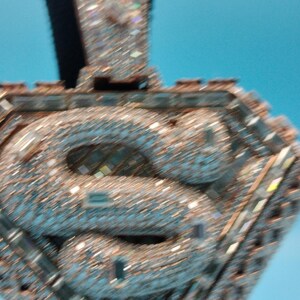 May include: A diamond-encrusted pendant with a stylized letter "S" at the center. The pendant is set with numerous small, sparkling stones, creating a shimmering effect. The design is geometric, with a rectangular top and a diamond-shaped frame.