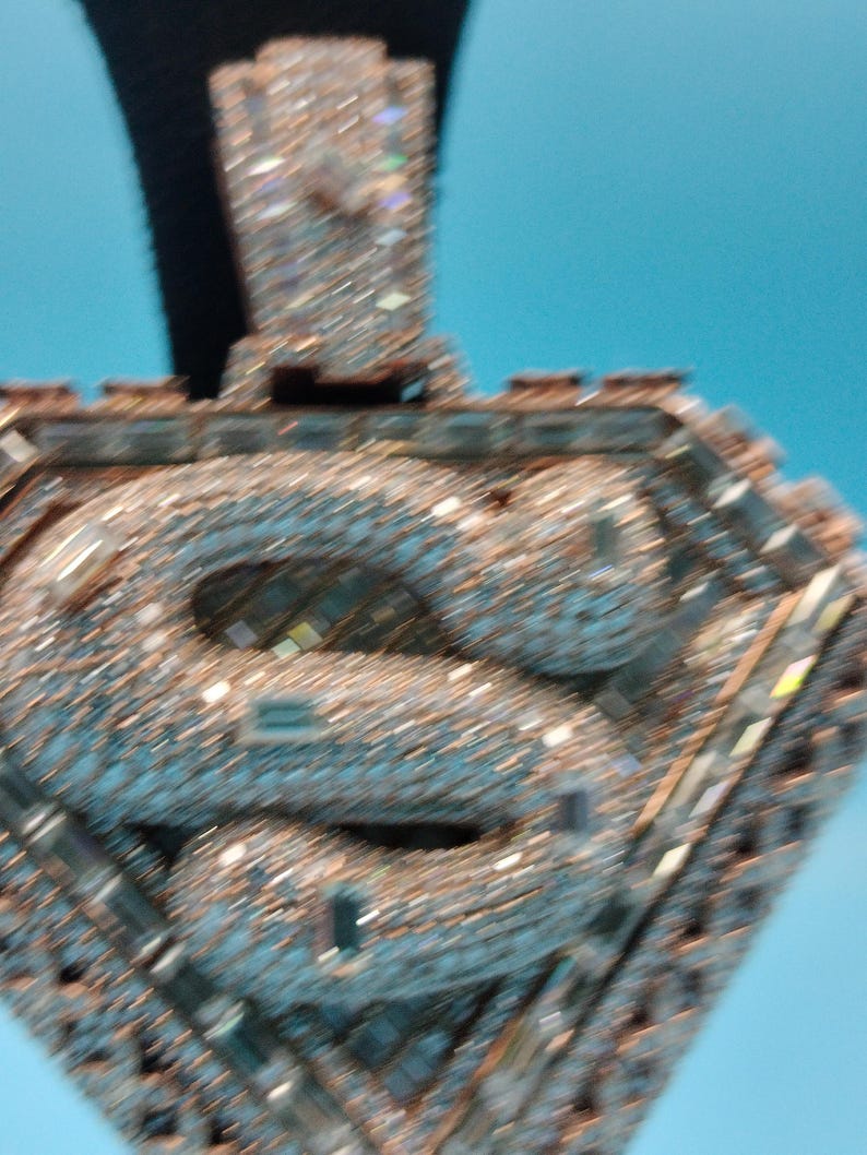 May include: A diamond-encrusted pendant with a stylized letter "S" at the center. The pendant is set with numerous small, sparkling stones, creating a shimmering effect. The design is geometric, with a rectangular top and a diamond-shaped frame.