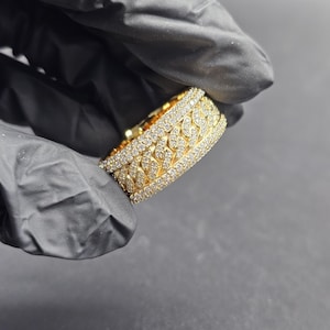 May include: A gold-colored ring with a chain link design, encrusted with small, clear gemstones. The ring is held against a dark background, showcasing its intricate details and sparkling appearance. The ring's design is a continuous pattern around the band.