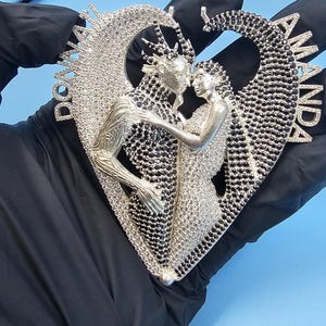 May include: A silver-toned pendant in the shape of a heart, featuring two figures embracing within a heart-shaped frame. The pendant is encrusted with small, sparkling stones, with the names "DONNA" and "AMANDA" on either side.