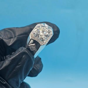 Peut inclure: Une bague en argent avec une grosse pierre précieuse ronde et transparente au centre, entourée de petites pierres. La bague a un design rectangulaire avec de petits détails carrés. Le fond est bleu clair uni.