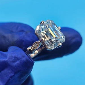 Peut inclure: Une bague en argent avec une grosse pierre précieuse claire de taille émeraude. La bague a un anneau délicat avec de petites pierres d'accent près de la pierre principale. Le fond est de couleur bleu uni.