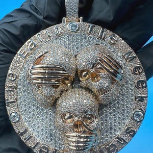 May include: A gold-toned pendant featuring three skull designs, each encrusted with small, sparkling stones. The pendant has a circular shape with the words "Earth to the Meek Shall Inherit" engraved around the edge. The skulls are in a see no evil, hear no evil, speak no evil pose.