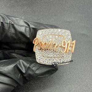 May include: A rose gold-toned ring featuring a rectangular face covered in small, clear, sparkling stones. The text "Psalm 91:1" is prominently displayed in raised, rose gold-toned script across the center of the stone-encrusted surface.