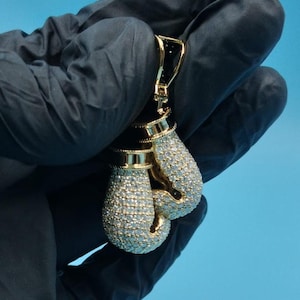 Peut inclure: Un pendentif doré en forme de gants de boxe. Les gants sont incrustés de petites pierres précieuses transparentes. Le haut du pendentif est noir avec des détails dorés. Le pendentif est présenté sur fond bleu.