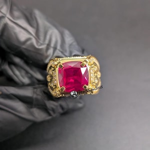 Puede incluir: Un anillo de oro con una gran piedra preciosa de color rojo intenso y corte cojín en el centro. La banda está decorada con intrincados trabajos en metal y pequeñas piedras claras. El texto Worldmine2005 es visible en la parte inferior derecha.