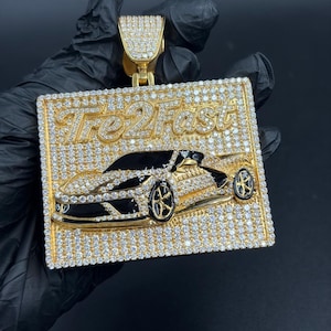 May include: A gold-colored pendant with the text "Tre2Fast" and a car design. The pendant is rectangular and encrusted with small, clear gemstones. The car is black and gold, with detailed wheels. The pendant is held by a gold bail.