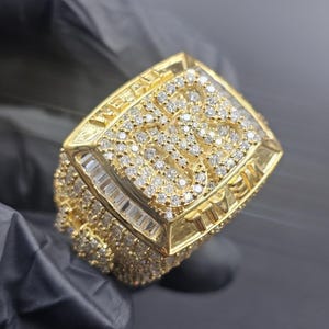 Puede incluir: Un anillo dorado con una parte superior rectangular, incrustado con numerosas piedras preciosas pequeñas y transparentes. El anillo tiene una banda gruesa y la palabra "WE ALL" es visible en el lateral. El anillo se sostiene sobre un fondo oscuro.