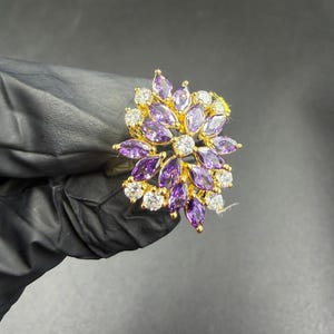 May include: A gold ring with a floral design. The ring is set with marquise-cut purple gemstones and round, clear gemstones. The gemstones are arranged in a flower-like pattern, set in a gold metal setting, creating a sparkling effect.