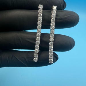 Peut inclure: Une paire de boucles d'oreilles linéaires. Chaque boucle d'oreille est ornée d'une série de pierres précieuses rondes et transparentes, disposées en ligne droite. Les boucles d'oreilles sont présentées sur fond bleu, tenues par une main gantée.