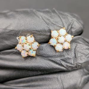May include: A pair of gold-tone stud earrings, each with a flower-shaped arrangement of white opal stones. The opals display iridescent colours. The earrings are presented against a dark backdrop.