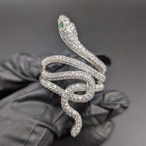 Może przedstawiać: Srebrny pierścionek w kształcie węża, inkrustowany małymi, przezroczystymi kamieniami szlachetnymi. Głowa węża ma dwa małe, zielone kamienie szlachetne jako oczy. Pierścionek owija się wokół palca.