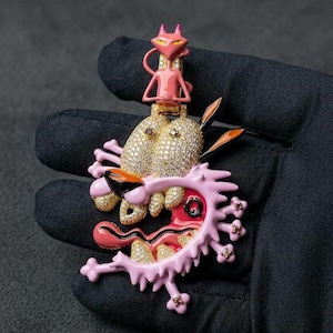 Peut inclure: Pendentif rose et doré représentant un chien de dessin animé avec une bouche ouverte et un chat rose. Le visage du chien est orné de pierres précieuses dorées et le chat est rose. Le pendentif est posé sur un fond noir.