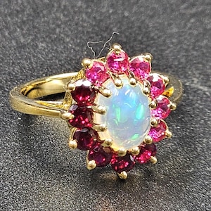 May include: A gold ring featuring a central oval opal gemstone surrounded by a halo of vibrant red gemstones. The ring's band is a simple, polished gold, and the setting showcases the gemstones' colors.