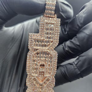 May include: Rose gold pendant encrusted with sparkling, rectangular-cut gemstones. The pendant is in the shape of the letters "J O Y" and is held by a gloved hand. The pendant has a polished finish and is set against a dark background.