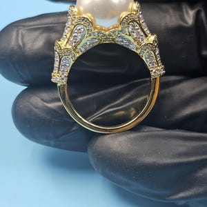 Peut inclure: Une bague dorée ornée d'une grosse perle blanche et ronde en son centre. La monture et le sertissage de la bague sont ornés de petites pierres claires et scintillantes. La bague est tenue sur un fond noir, avec une surface bleu clair visible.