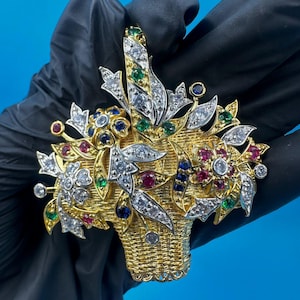 以下が含まれることがあります： 宝石の花の配置が施された、金色のバスケットブローチ。ブローチは、ダイヤモンド、ルビー、エメラルド、サファイアで飾られた葉と花が付いた織りバスケットのデザインが特徴です。ブローチは青い背景にあります。