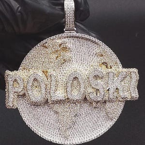 May include: A large, round pendant with the word "POLOSKI" in bold, diamond-encrusted letters. The pendant is gold-toned and covered in small, sparkling stones, with a detailed map design in the background. A diamond-studded bail is at the top.