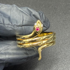 Może przedstawiać: Złoty pierścionek w kształcie węża z rubinem i dwoma przezroczystymi kamieniami na głowie. Pierścionek owija się wokół palca, z teksturowanym wzorem na obrączce. Ogon węża jest zakrzywiony.