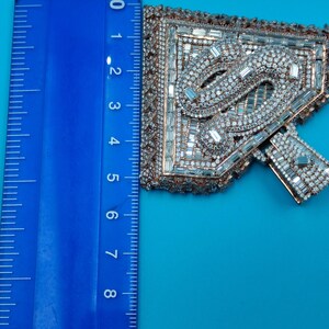 May include: An ornate, jeweled applique with a geometric design, featuring clear and silver-toned crystals. The applique is approximately 4 inches long, with a copper-colored border, set against a blue background.