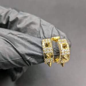 May include: Gold-toned hoop earrings with a studded design. The earrings feature small, pointed studs and are encrusted with small, clear gemstones. The earrings are displayed against a dark background.