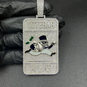 May include: A rectangular pendant with a diamond-like encrusted surface. The pendant features a cartoon character in a tuxedo, running with a money bag and green notes. The word "RICH BAG" is at the top of the pendant.