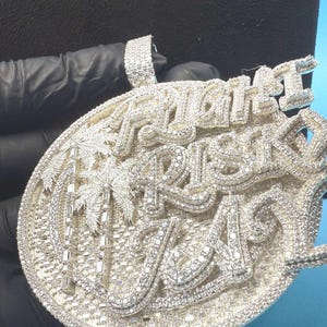 May include: A silver pendant with the words "RIGHT RISK A" in a stylized font. The pendant features a palm tree design and is encrusted with small, sparkling stones. The pendant has a loop at the top for a chain.