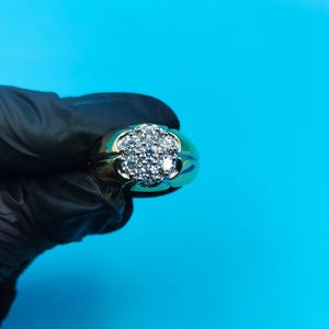 Puede incluir: Un anillo dorado con un grupo de pequeñas piedras preciosas transparentes dispuestas en un patrón floral. El anillo tiene una banda ancha y una parte superior redondeada, sobre un fondo azul brillante.