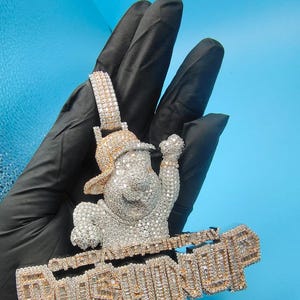 May include: A diamond-encrusted pendant featuring a cartoon character wearing a hat and raising a hand. The pendant has the word "PUSHINP" in a gold-colored, block-letter design. The pendant is held against a black surface.