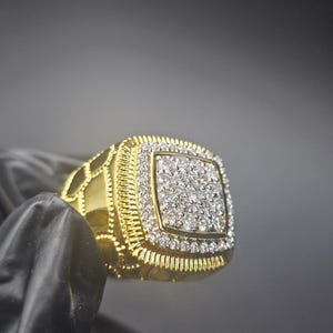Puede incluir: Un anillo dorado con una cara cuadrada, incrustado con pequeñas piedras preciosas transparentes. La banda del anillo presenta un diseño texturizado, y la cara está bordeada por una fila de piedras más pequeñas. El anillo se muestra sobre un fondo oscuro.