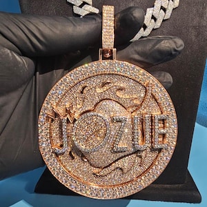 May include: A large, round pendant necklace with the name "OZUE" spelled out in sparkling, diamond-like stones. The pendant has a rose gold tone and is encrusted with many small, clear stones. A matching chain is visible in the background.
