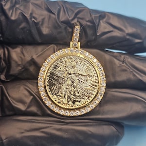 Peut inclure: Un pendentif doré avec un relief détaillé d'une figure, entouré d'un anneau de pierres précieuses transparentes. Le pendentif a une boucle en haut, également ornée de pierres précieuses. Le fond est bleu clair uni.