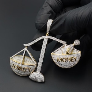 以下が含まれることがあります： 「LOYALTY」と「MONEY」の文字が両側に刻まれた、金と銀の天秤の形をしたペンダント。ペンダントには、小さな輝く石がちりばめられています。