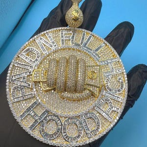 May include: A large, round pendant with a gold and silver design, encrusted with numerous small, sparkling stones. The pendant features the words "HARD WORK" and "RICH HOOD" in a bold font, along with a fist and money bag design.