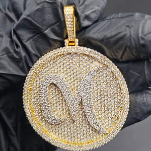 May include: A gold-coloured pendant with a large, circular design. The pendant is covered in many small, sparkling stones. The letter "M" is in the centre, also decorated with smaller stones. The bail is also covered in stones.