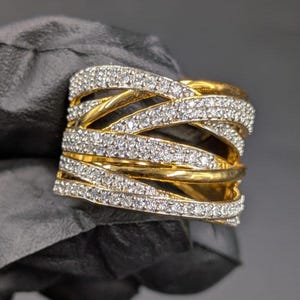 May include: A gold ring with a woven design. The ring has multiple bands, some set with small, sparkling white gemstones. The ring's design creates a layered, textured appearance, showcasing a blend of gold and white.