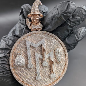 May include: A large, round pendant with a rose gold and silver design. The pendant features a detailed witch figure at the top and the letters "MMM" in the center, all encrusted with sparkling stones. A small bag of money and a coffin are also visible.