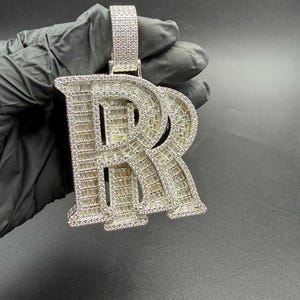 May include: A silver-colored pendant in the shape of the letter "R", encrusted with small, sparkling stones. The pendant has a rectangular bale and is held by a gloved hand against a dark background. The stones are arranged to give the pendant a brilliant shine.