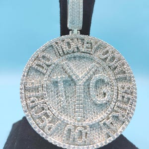 May include: A round, silver-colored pendant with the text "DO MONEY GOOD PEOPLE" encircling the outer edge. The center features the letters "TYG". The pendant is encrusted with small, sparkling stones and has a matching bail.