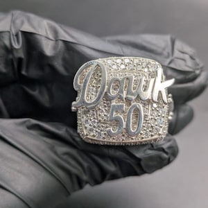 May include: A silver ring with the word "Dawk" in cursive and the number "50" in a block font. The ring is encrusted with small, clear stones. The ring is shown against a black background.
