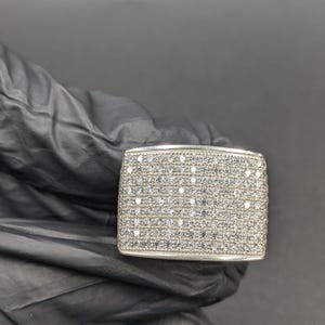 May include: A rectangular silver ring with a surface covered in small, clear stones. The ring has a rounded top and a smooth, polished finish. The ring is shown against a black background.