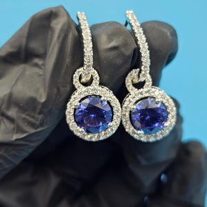 May include: A pair of drop earrings with large, round, deep blue gemstones encircled by smaller, clear, sparkling stones. The earrings have a silver-coloured metal setting and a lever back closure. The background is a solid light blue.